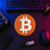 Terracotta orange circular Bitcoin crypto logo sign displayed flat on a neon-lit trader desk beside a keyboard, hardware wallet, and technical equipment.