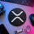 Black and white circular XRP crypto logo sign displayed flat on a neon-lit trader desk beside a keyboard, hardware wallet, and technical equipment.