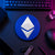 Cobalt blue circular Ethereum crypto logo sign displayed flat on a neon-lit trader desk beside a keyboard, hardware wallet, and technical equipment.