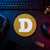 Gold circular Dogecoin crypto logo sign displayed flat on a neon-lit trader desk beside a keyboard, hardware wallet, and technical equipment.