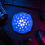Blue circular Cardano crypto logo sign displayed flat on a neon-lit trader desk beside a keyboard, hardware wallet, and technical equipment.