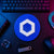 Blue circular Chainlink crypto logo sign displayed flat on a neon-lit trader desk beside a keyboard, hardware wallet, and technical equipment.