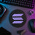 Black and purple circular Solana crypto logo sign displayed flat on a neon-lit trader desk beside a keyboard, hardware wallet, and technical equipment.